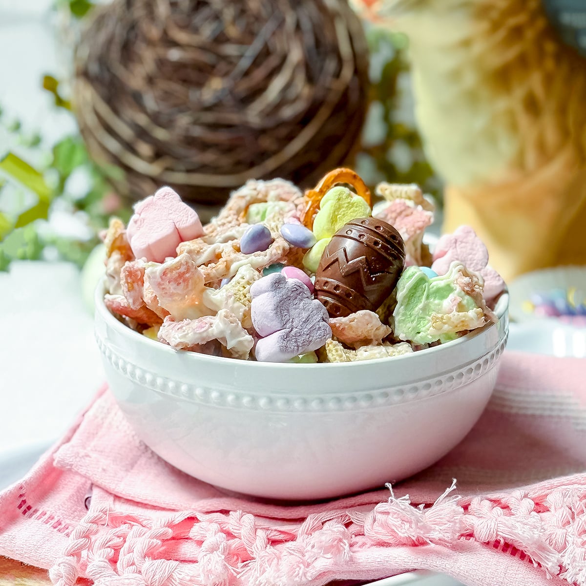 A white bowl filled with colorful Easter Snack Mix—cereal, heart-shaped marshmallows, and a chocolate egg—rests on a pink napkin, with a nest and giraffe figurine in the background.