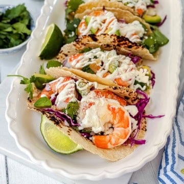 Three shrimp tacos with cabbage, greens, and creamy sauce are served on a white tray with lime wedges and cilantro-an irresistible shrimp tacos recipe for any occasion.