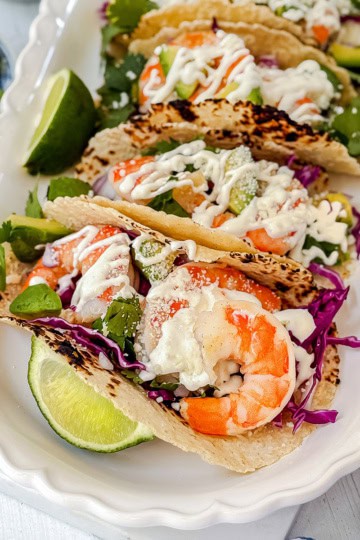 Three shrimp tacos with cabbage, greens, and creamy sauce are served on a white tray with lime wedges and cilantro-an irresistible shrimp tacos recipe for any occasion.