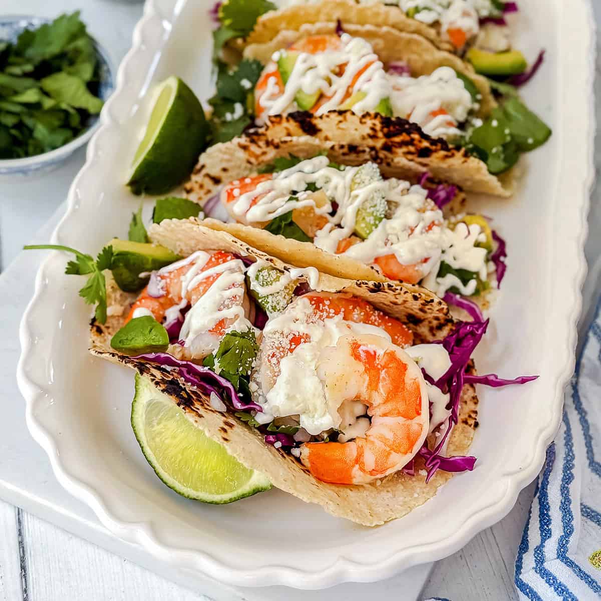 Three shrimp tacos with cabbage, greens, and creamy sauce are served on a white tray with lime wedges and cilantro-an irresistible shrimp tacos recipe for any occasion.