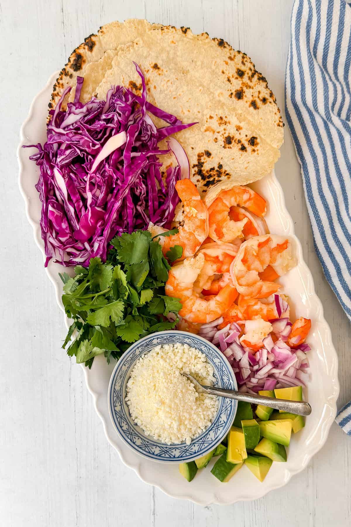 A white platter with corn tortillas, shredded purple cabbage, cooked shrimp, chopped red onion, cilantro, diced avocado, and a bowl of crumbled cheese, next to a blue-striped cloth on a white surface.