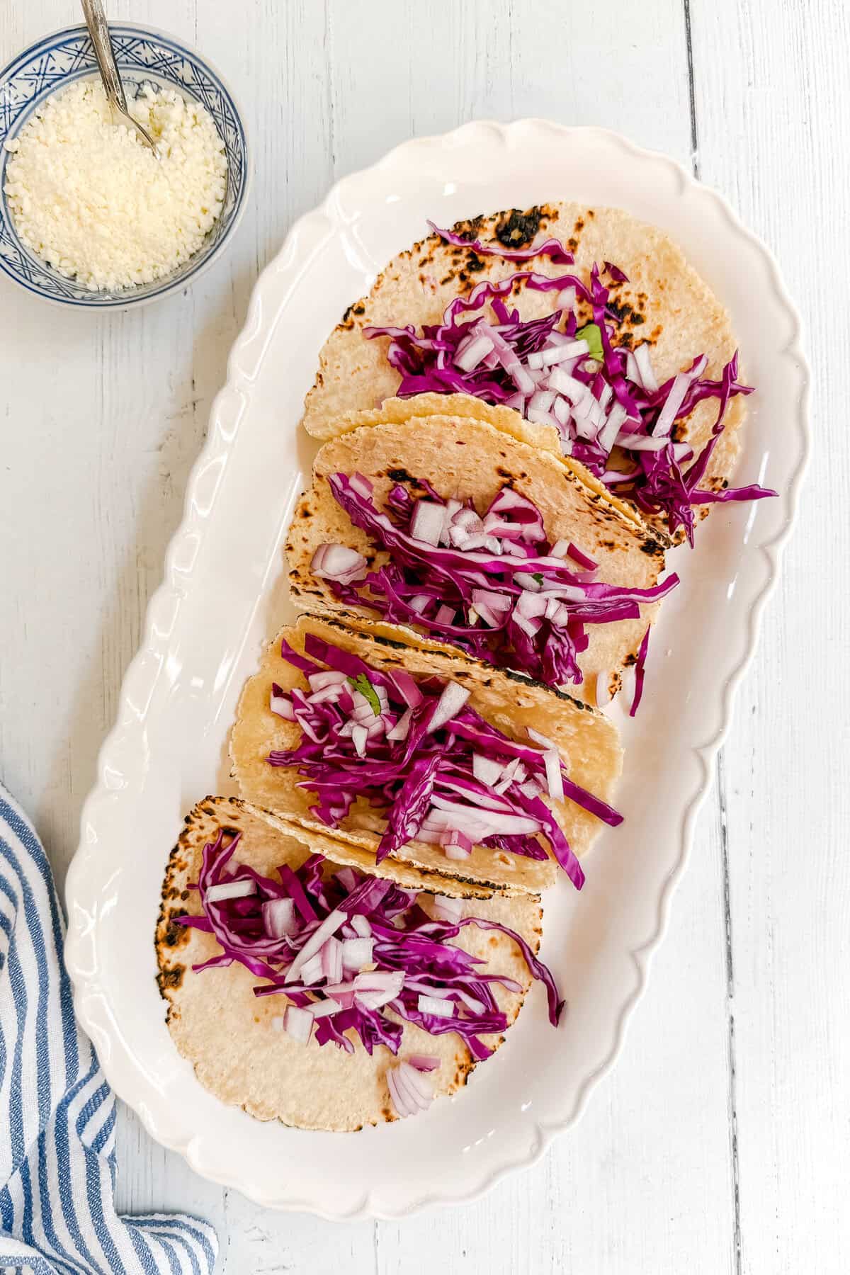 Three soft tacos filled with shredded purple cabbage and chopped onions are arranged on a white oval platter. A small bowl of grated cheese sits nearby on a white wooden surface.