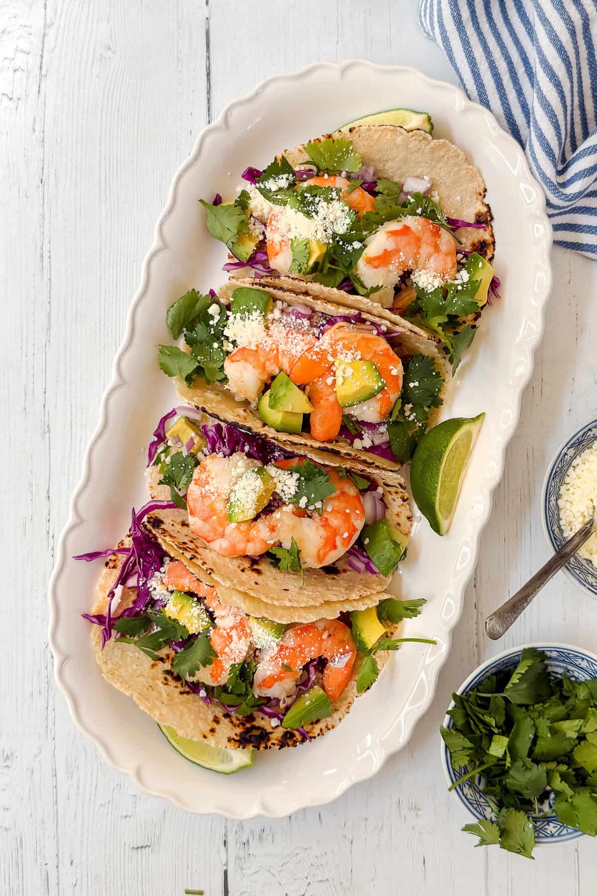 Three shrimp tacos filled with shrimp, avocado, purple cabbage, cilantro, and crumbled cheese are arranged on a white platter with lime wedges. A bowl of cilantro, cheese, and a striped napkin are nearby.