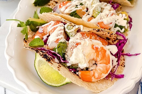 Four shrimp tacos on toasted corn tortillas are arranged on a white platter. They are topped with shredded cabbage, avocado slices, fresh cilantro, and creamy white sauce, with lime wedges on the side. Small bowls of toppings are in the background.