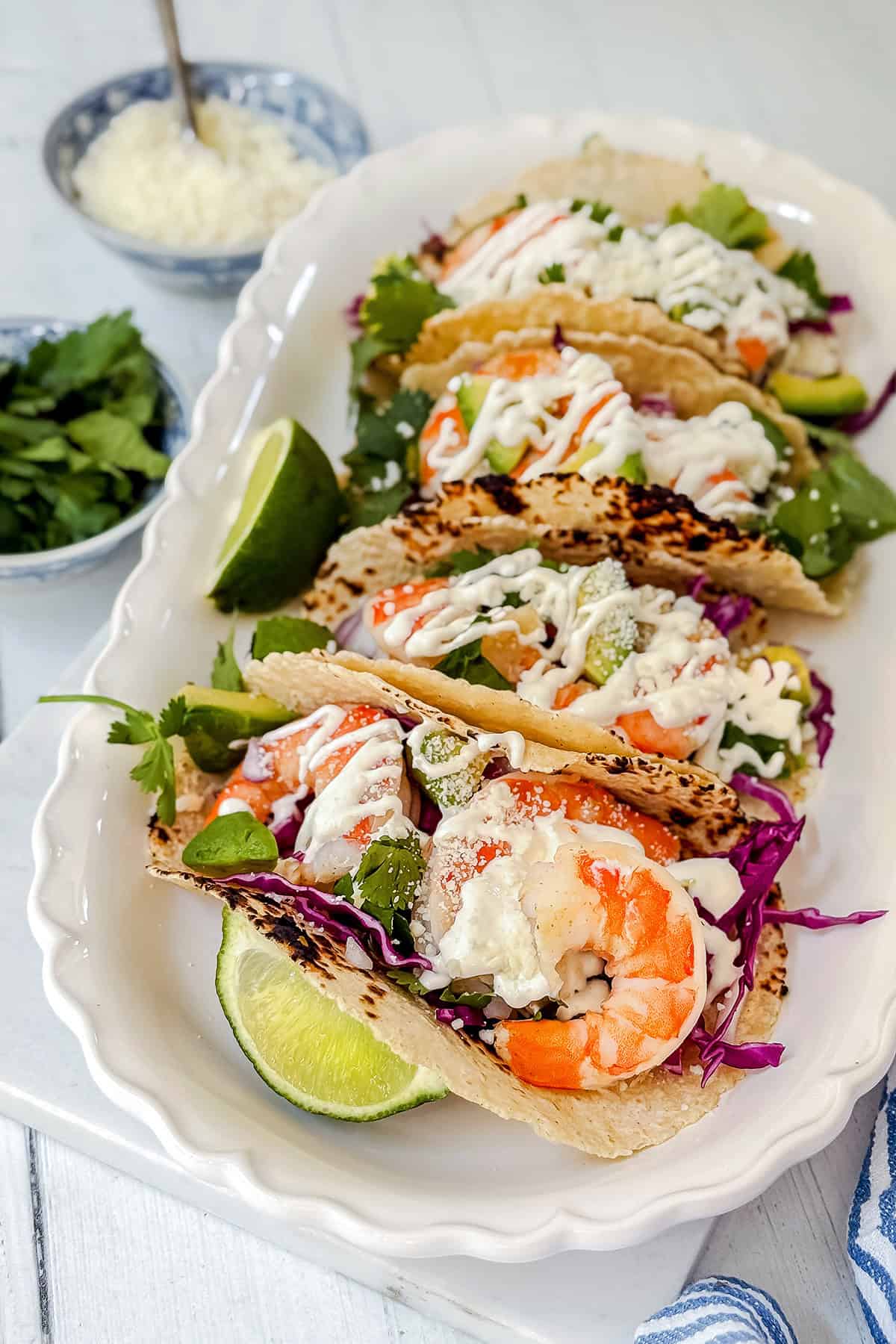 A platter of shrimp tacos, crafted from a flavorful shrimp tacos recipe, with shredded cabbage, avocado, cilantro, lime wedges, and a drizzle of white sauce, served on a white dish.
