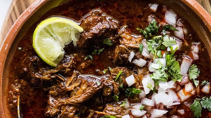 A bowl of spicy beef stew topped with chopped onions, cilantro, and a lime wedge, with a spoon inside. Warm tortillas and a bowl of chopped herbs are nearby on a wooden surface.