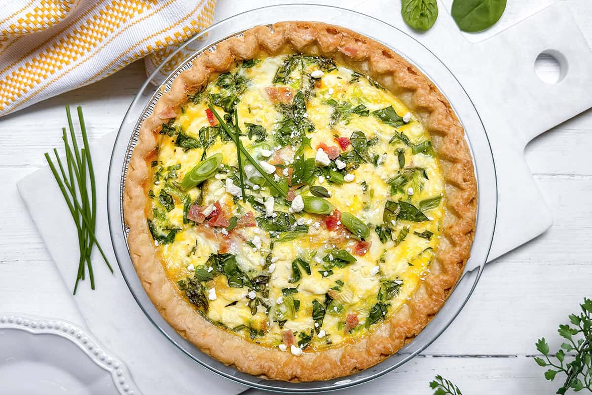 A baked asparagus quiche with a golden crust, topped with fresh herbs, sits on a white cutting board next to a yellow-striped towel and scattered greens.