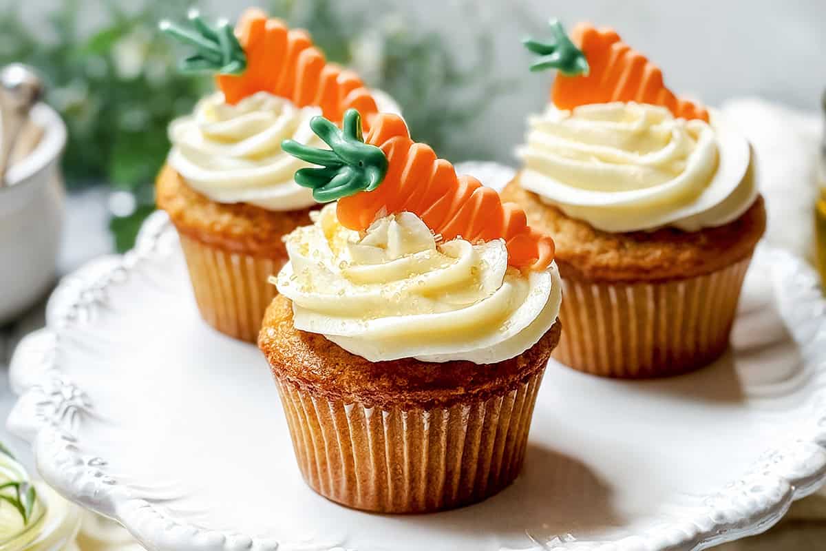 Three carrot cake cupcakes with swirled white frosting, topped with decorative carrot-shaped candies, are displayed on a white plate.