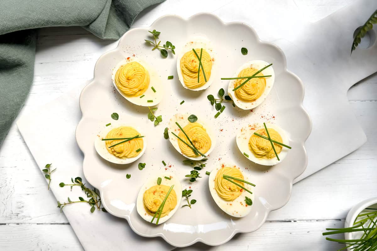 A white scalloped plate holds eight deviled eggs, each garnished with chives and a sprinkle of paprika. This tantalizing deviled eggs recipe is presented on a light wooden table with some scattered greenery. A green napkin is partially visible in the top left corner.