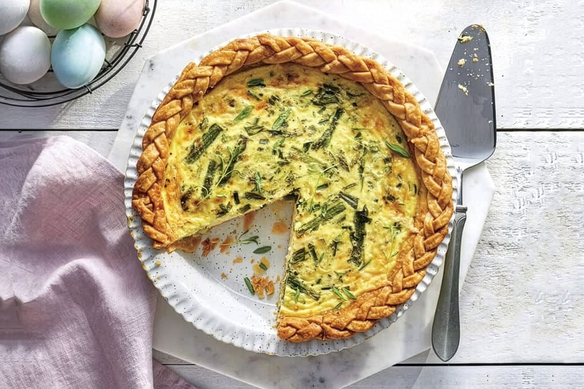 A quiche with a braided crust sits on a white plate, missing one slice&mdash;perfect for Easter Brunch. A pie server rests nearby, with pastel-colored eggs in a bowl completing the festive scene.