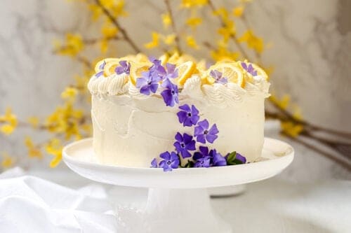A white frosted lemon cake recipe, decorated with lemon slices and purple flowers, sits on a white cake stand, with yellow flowers blurred in the background.