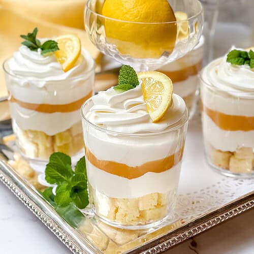 Four glass cups filled with a layered lemon dessert trifle are displayed on a silver tray. Each cup contains cake pieces, lemon curd, and whipped cream, topped with a lemon slice and mint. A whole lemon sits in the background next to a yellow cloth.