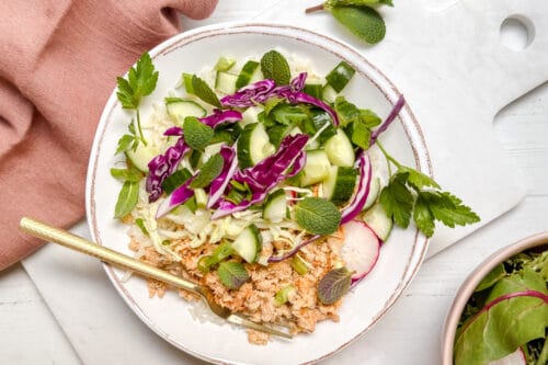 A vibrant salmon bowl featuring minced meat topped with sliced cucumber, purple cabbage, fresh mint, parsley, and radish slices. A gold fork rests inside the bowl, with a pink napkin partially visible on the side.