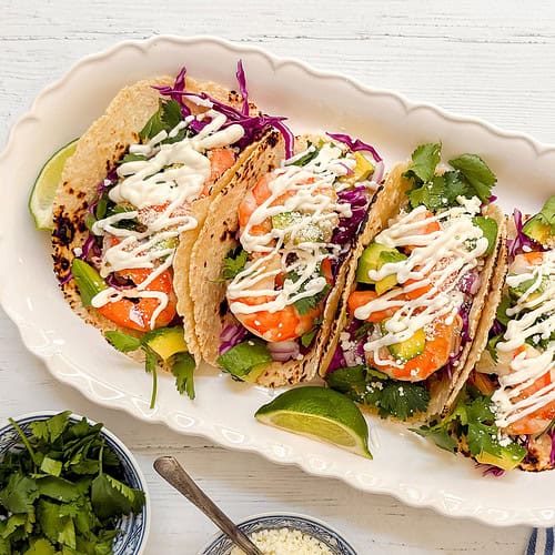 A white dish holds four tacos filled with shrimp, avocado, purple cabbage, cilantro, and cheese, topped with a drizzle of creamy sauce. Lime wedges, fresh cilantro, and cheese are on the side.