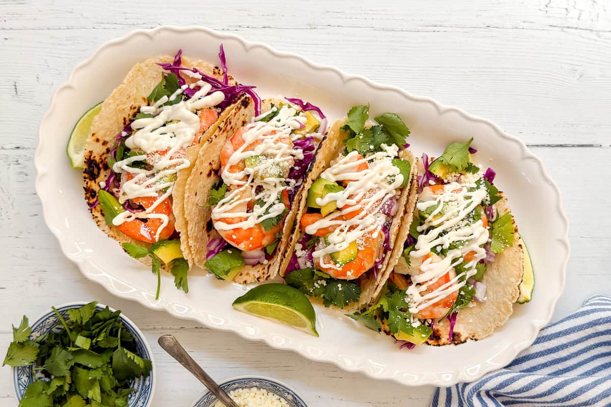 A white dish holds four tacos filled with shrimp, avocado, purple cabbage, cilantro, and cheese, topped with a drizzle of creamy sauce. Lime wedges, fresh cilantro, and cheese are on the side.