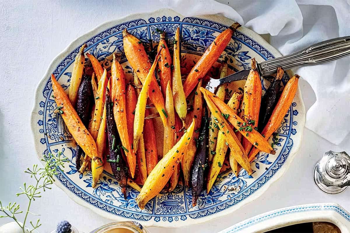 A blue and white platter holds roasted carrot sticks garnished with herbs, displayed on a white tablecloth with silverware beside the dish-an elegant choice for Easter side dishes.