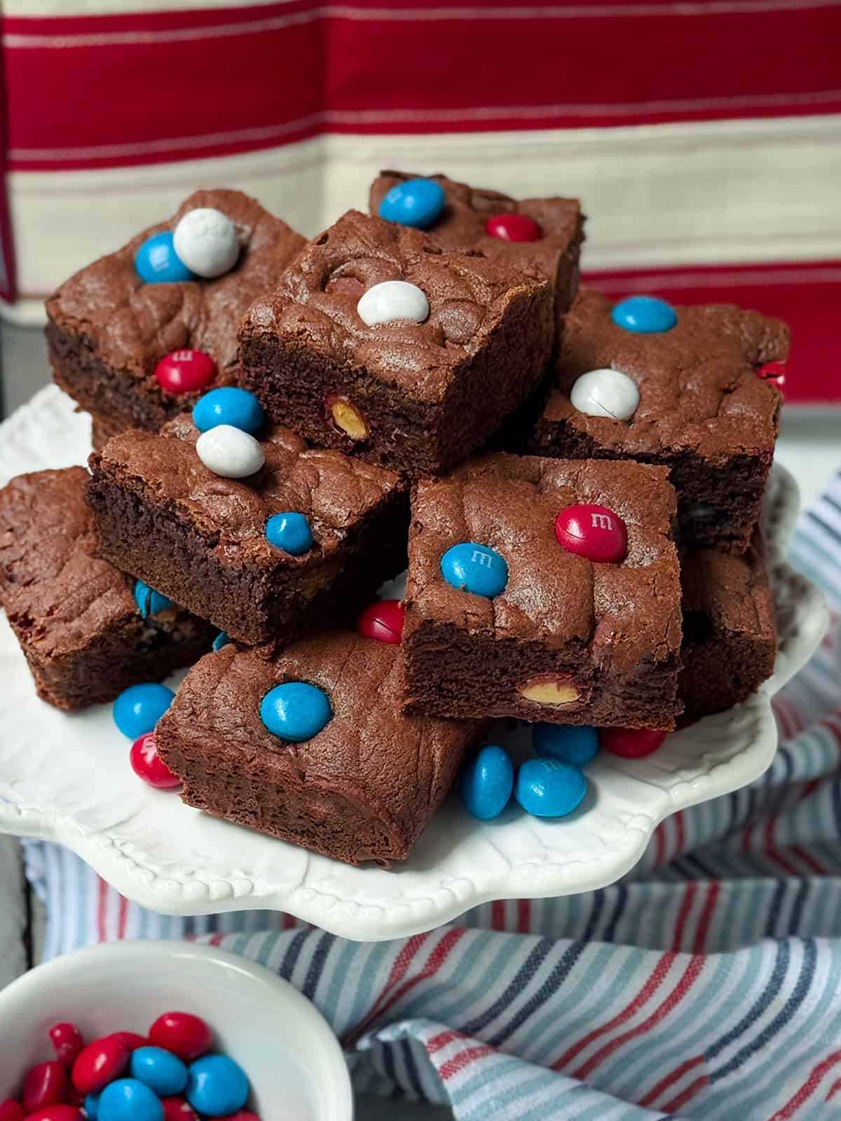 A plate of chocolate brownies, reminiscent of festive M&M Cookie Bars, topped with red, white, and blue candy-coated chocolates. More colorful candies fill a bowl beside a striped cloth in the background.