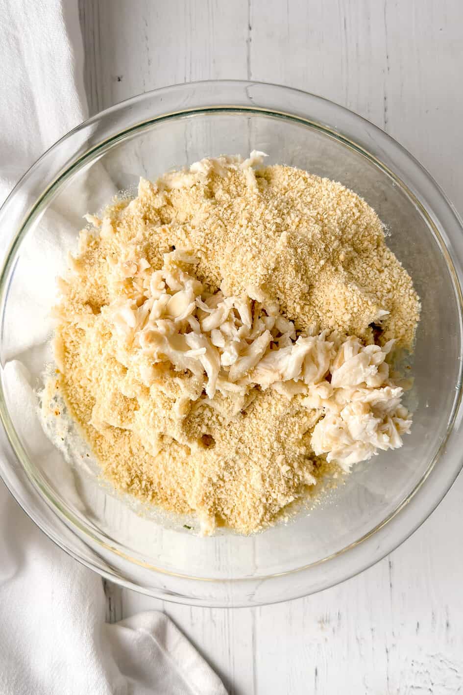 A glass bowl on a white wooden surface holds crab meat and breadcrumbs, ready to be mixed for Maryland crab cakes. A white cloth with a blue stripe is partially visible beside the bowl.