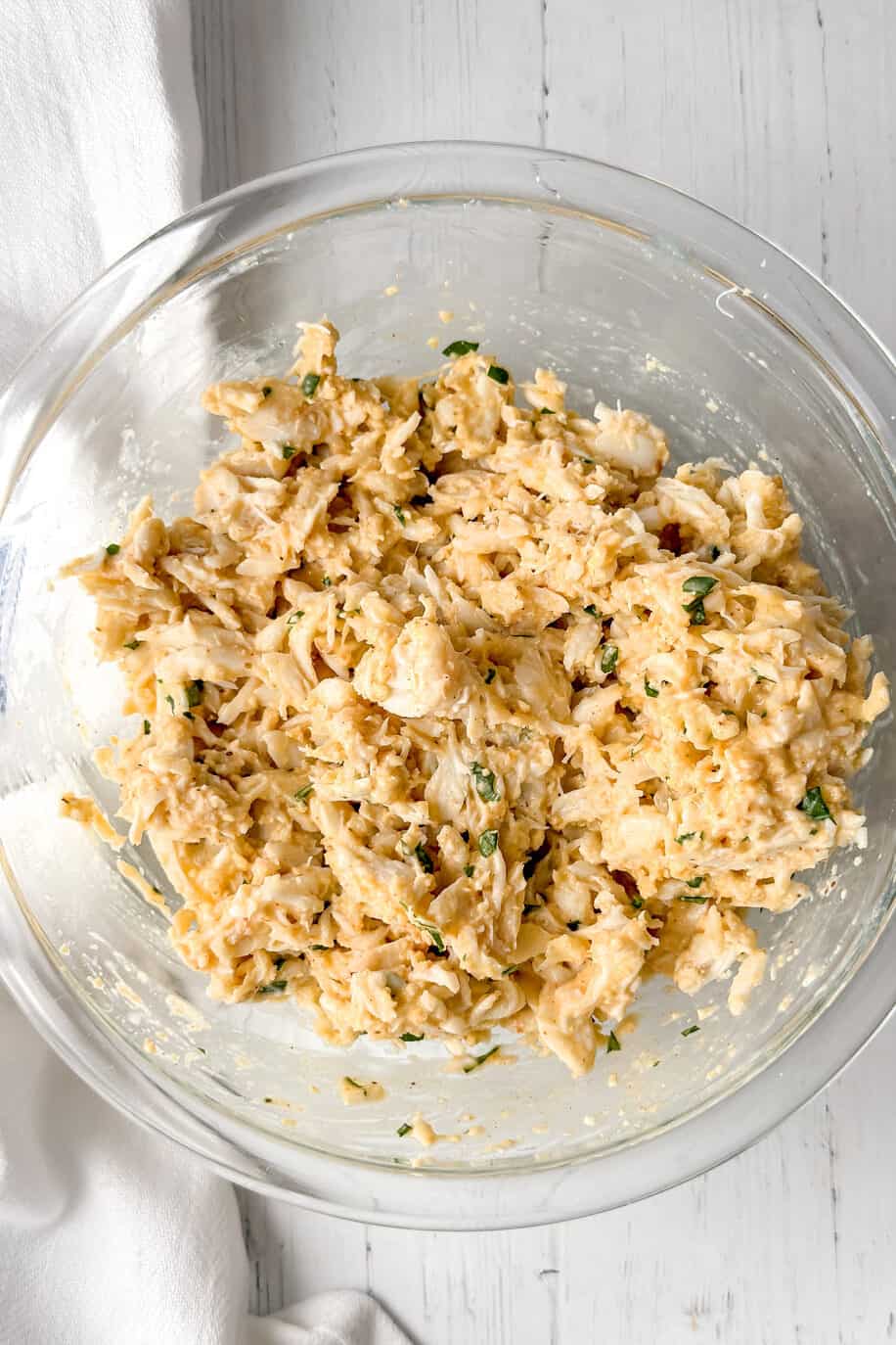 A glass bowl filled with a creamy crab salad mixture sits on a white wooden surface, reminiscent of the fresh flavors found in Maryland crab cakes, next to a folded white towel with a blue stripe.