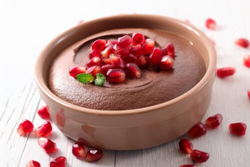 A round dish of creamy chocolate mousse topped with pomegranate seeds and a small mint leaf, with more pomegranate seeds scattered on a white wooden surface.