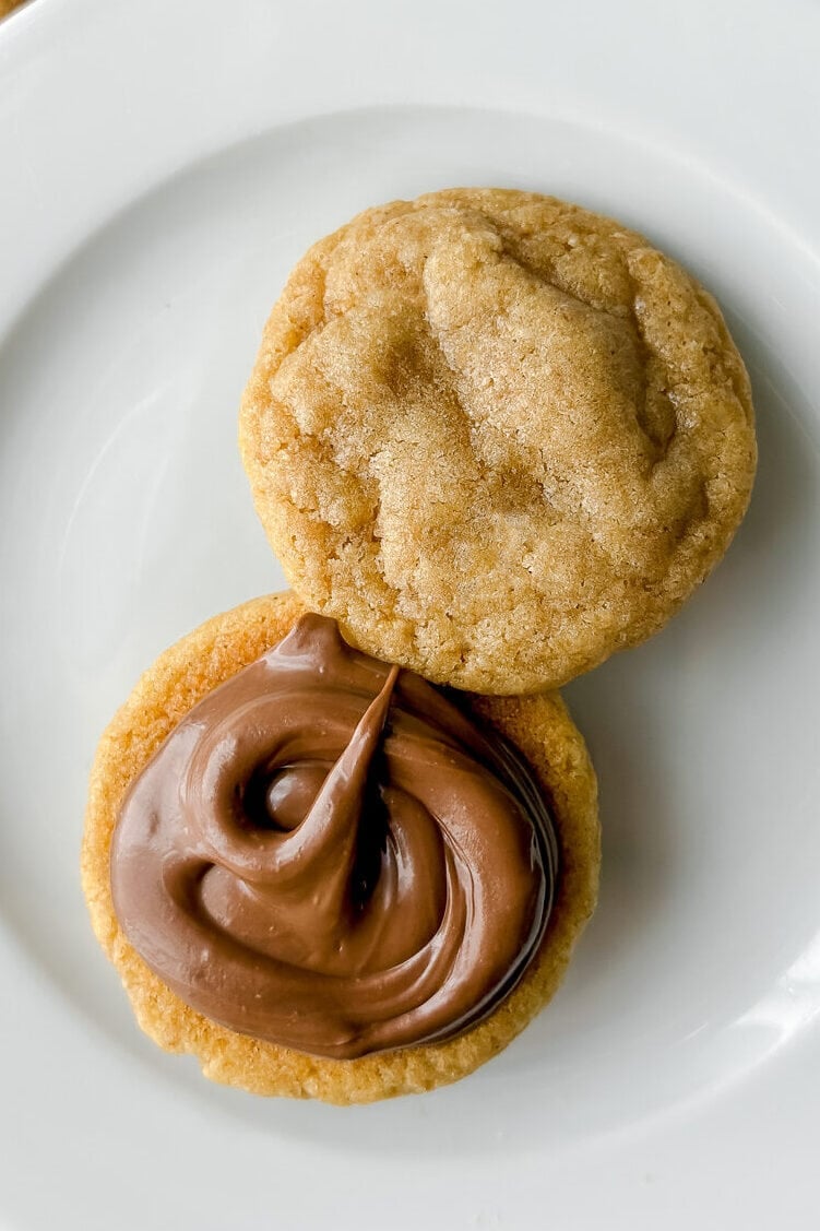 A white plate holds two round graham cookies, one topped with a swirl of chocolate spread. A striped cloth napkin and a square graham cracker are partly visible beside the plate.