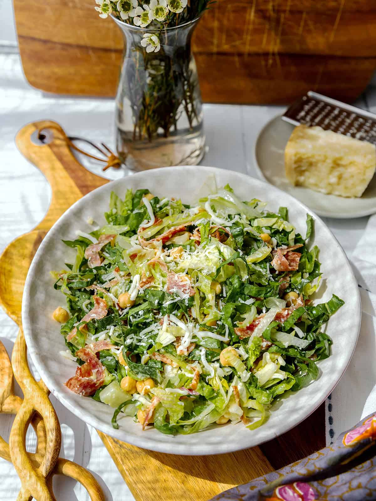A bowl of La Scala chopped salad with lettuce, chickpeas, tomatoes, grated cheese, and dressing. A block of cheese, grater, and flowers are in the background.