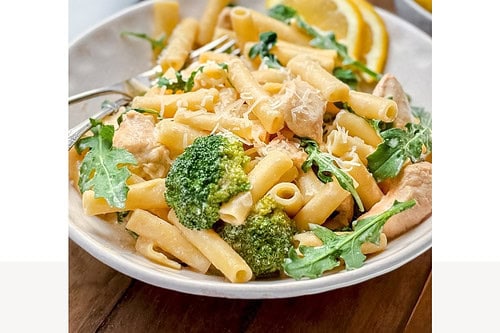 A bowl of creamy chicken pasta with broccoli and arugula, topped with grated cheese, sits on a wooden table with lemon slices and bread in the background. Text reads: "Easy One Pot Creamy Chicken Pasta. Dinner in 30 minutes!.