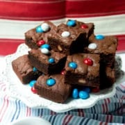 A plate of chocolate brownies topped with red, white, and blue candy-coated chocolates, reminiscent of festive M&M Cookie Bars, is displayed on a white plate with a patriotic red and white background.