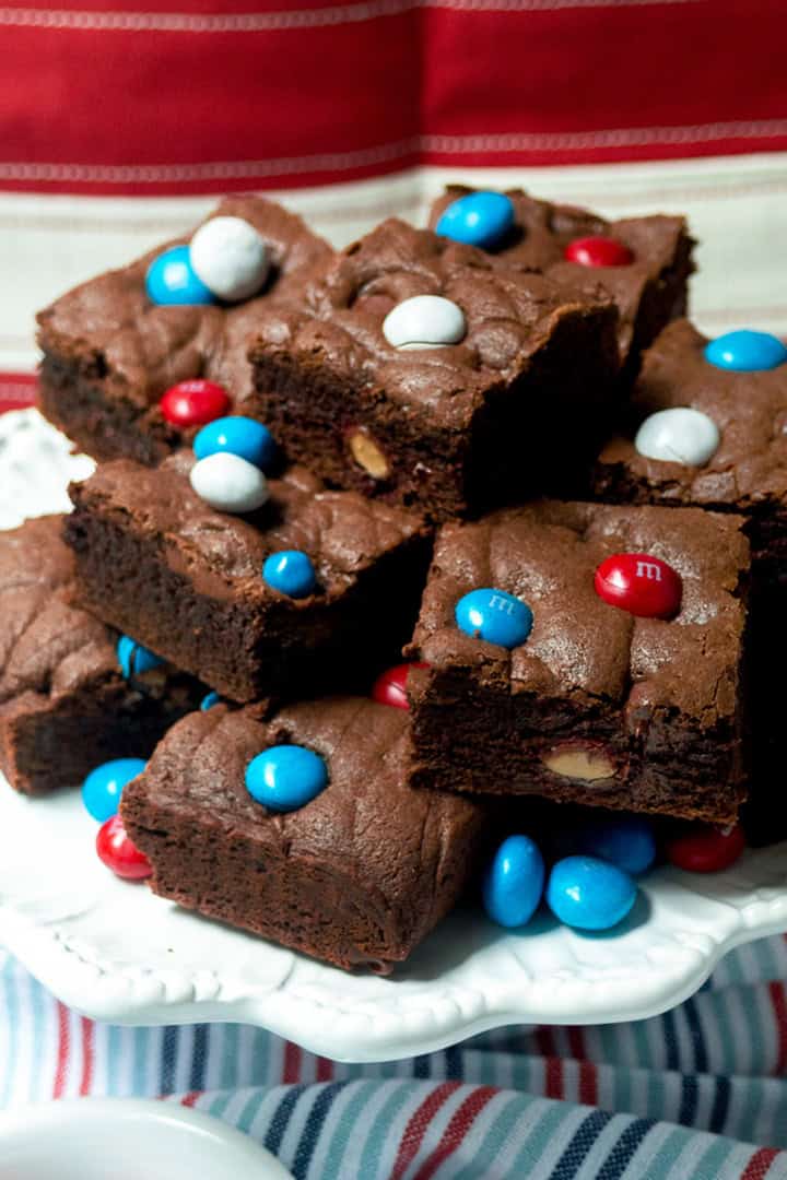 A plate of chocolate brownies topped with red, white, and blue candy-coated chocolates, reminiscent of festive M&M Cookie Bars, is displayed on a white plate with a patriotic red and white background.