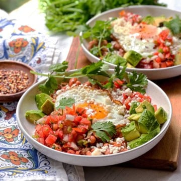 Two bowls of huevos rancheros with fried eggs, avocado, fresh salsa, beans, cheese, and cilantro.