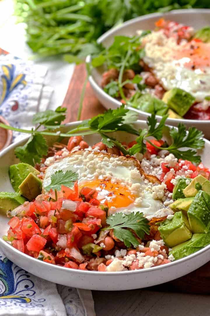 Two bowls of huevos rancheros with fried eggs, avocado, fresh salsa, beans, cheese, and cilantro.