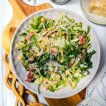 A bowl of la scala chopped salad with lettuce, chickpeas, shredded cheese, and slices of cured meat sits on a wooden board.