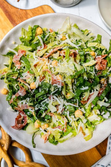 A bowl of la scala chopped salad with lettuce, chickpeas, shredded cheese, and slices of cured meat sits on a wooden board.