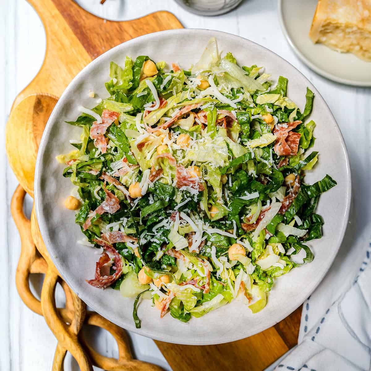 A bowl of la scala chopped salad with lettuce, chickpeas, shredded cheese, and slices of cured meat sits on a wooden board.