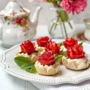 Six strawberry tarts topped with cream and strawberry slices arranged like roses, garnished with mint leaves, served on a white plate with a floral teapot and vase in the background.
