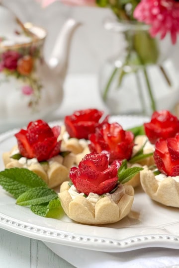 Six strawberry tarts topped with cream and strawberry slices arranged like roses, garnished with mint leaves, served on a white plate with a floral teapot and vase in the background.