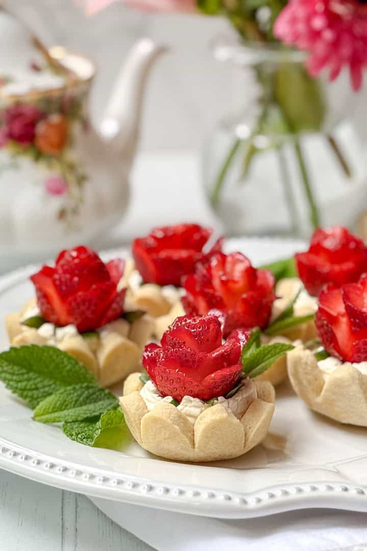 Six strawberry tarts topped with cream and strawberry slices arranged like roses, garnished with mint leaves, served on a white plate with a floral teapot and vase in the background.