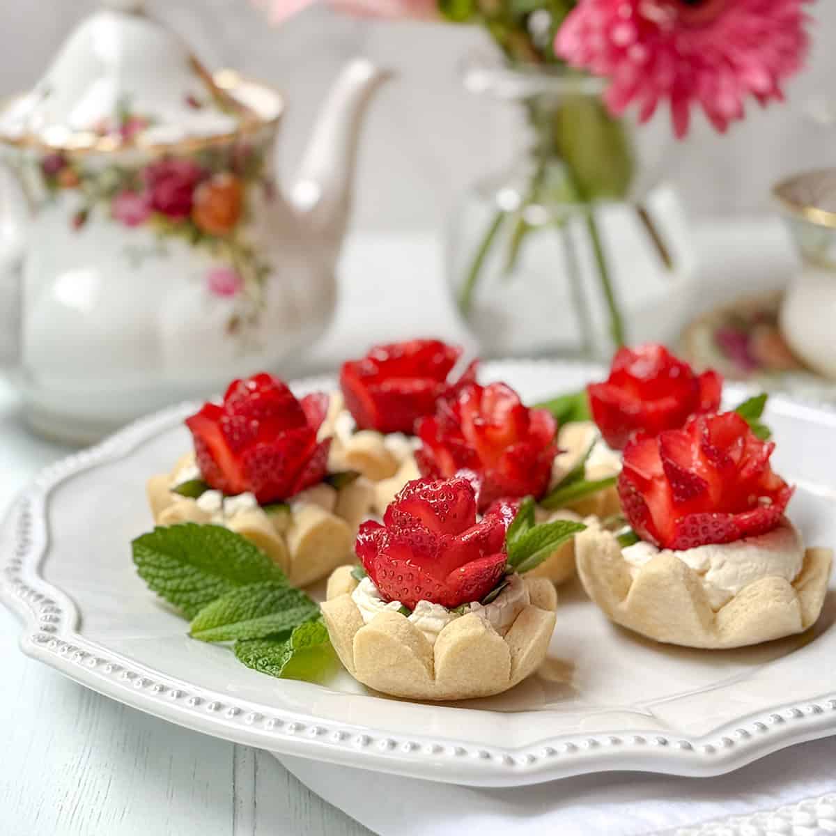 Six strawberry tarts topped with cream and strawberry slices arranged like roses, garnished with mint leaves, served on a white plate with a floral teapot and vase in the background.