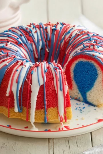 A festive bundt cake with red, white, and blue layers and icing, topped with sprinkles, sits on a plate with a slice removed to reveal its colorful interior-perfect for Memorial Day Desserts.