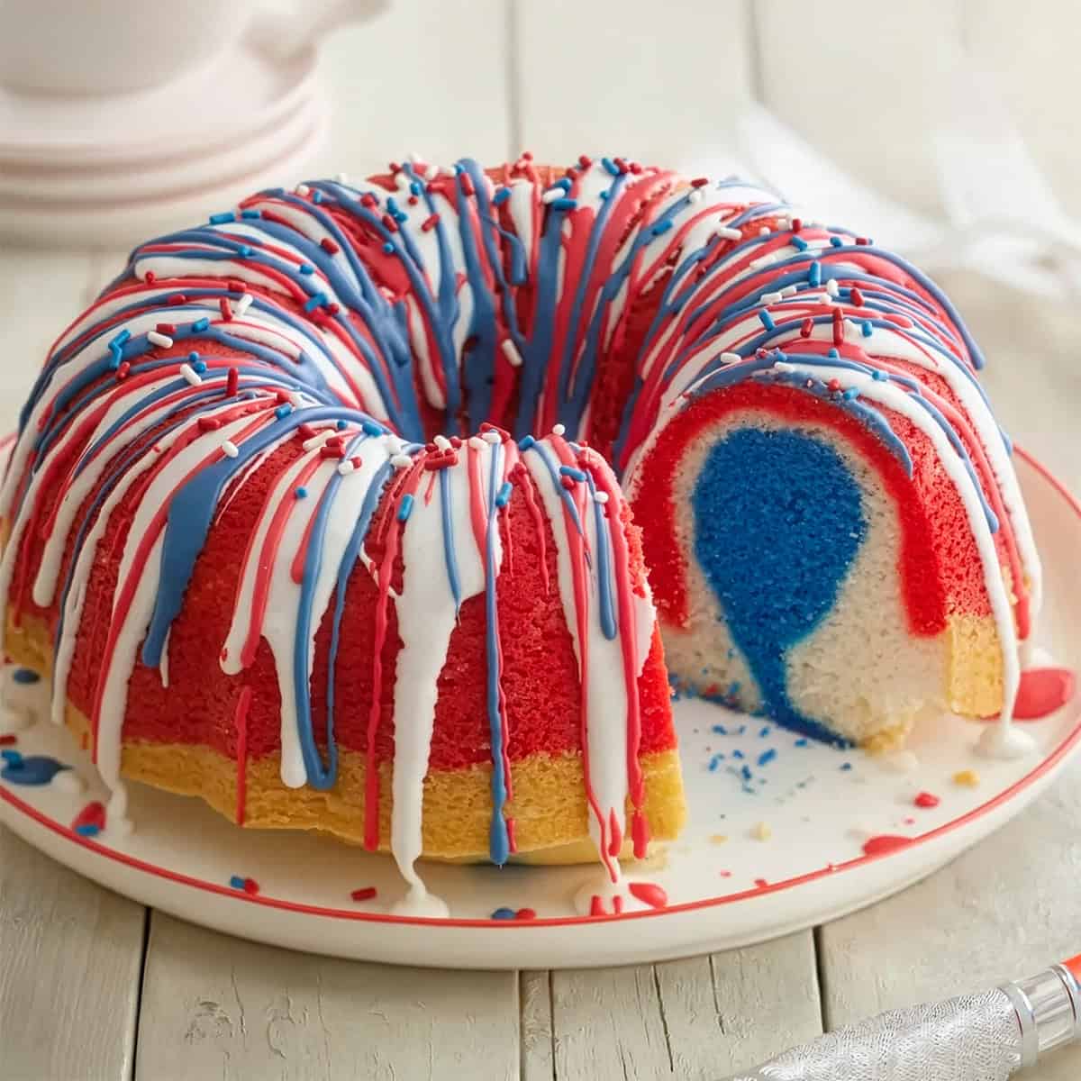 A festive bundt cake with red, white, and blue layers and icing, topped with sprinkles, sits on a plate with a slice removed to reveal its colorful interior—perfect for Memorial Day Desserts.