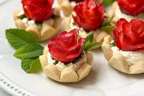 A white plate with small strawberry tarts, each topped with rose-shaped sliced strawberries and garnished with mint. The text “Strawberry Tarts” appears above the tarts.
