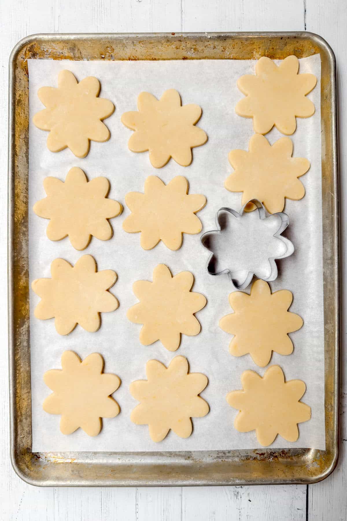 A baking sheet lined with parchment paper holds twelve unbaked flower-shaped cookies, perfect for pairing with strawberry tarts. A metal flower-shaped cookie cutter rests on one cookie cutout atop a white wooden surface.