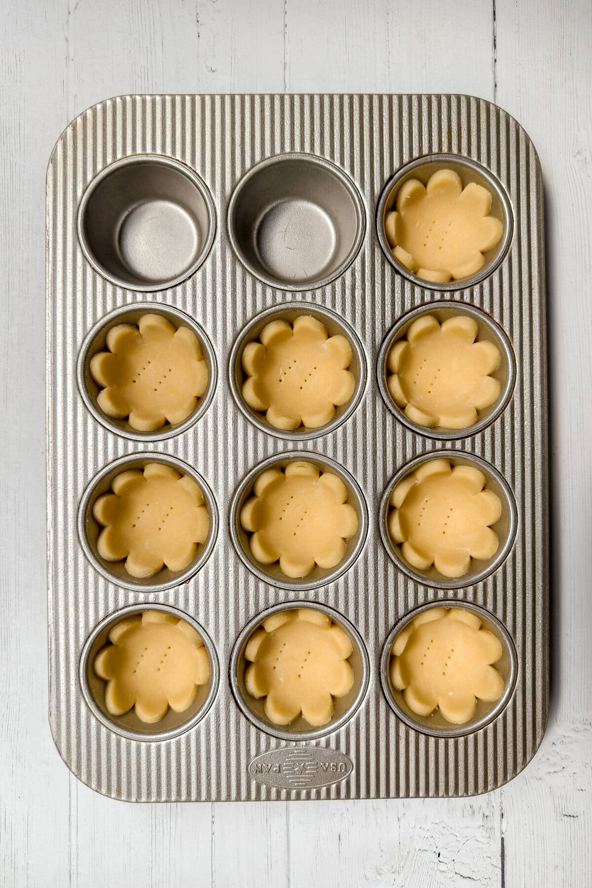 A metal muffin tin holds ten unbaked flower-shaped tart shells, ready for delicious strawberry tarts; two cups are empty. The tin rests on a white wooden surface.