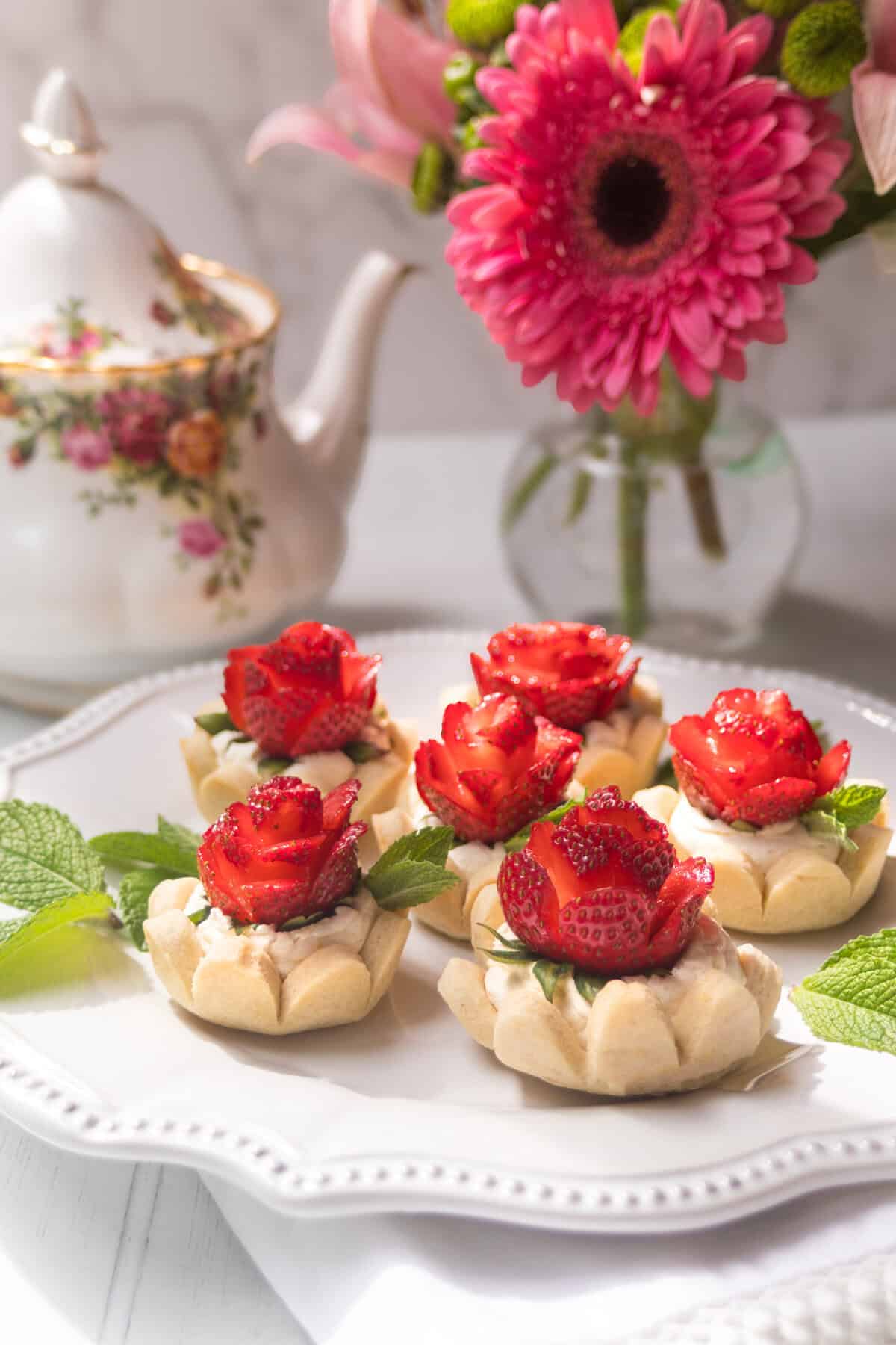 Six rose-shaped strawberry tarts topped with strawberry roses and mint leaves are arranged on a white plate. A floral teapot and a vase with pink flowers in the background create a bright and elegant tea setting.