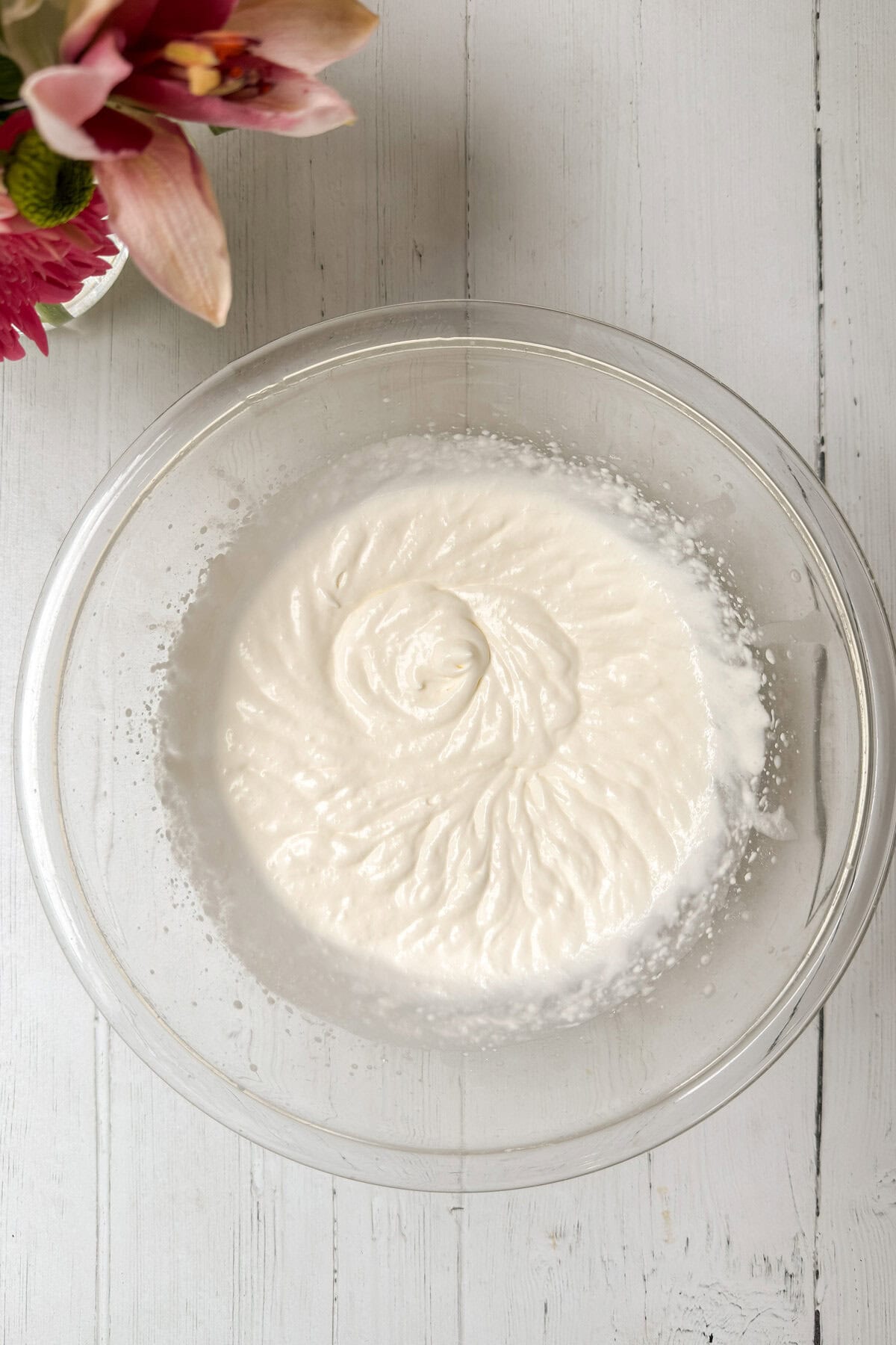 A clear glass bowl filled with whipped coconut cream sits on a white wooden surface, ready to top freshly baked strawberry tarts. Pink flowers in the upper left corner add a touch of color to the scene.