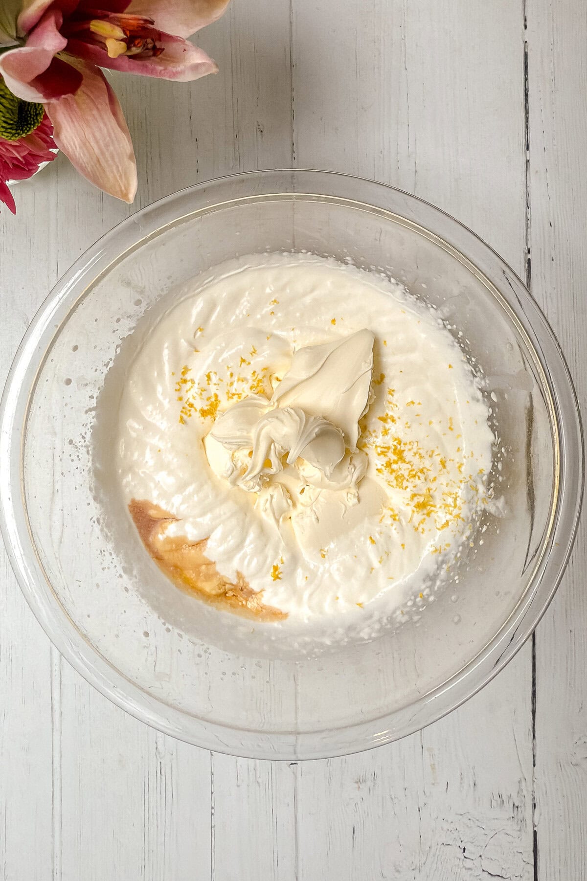 A glass bowl filled with a mixture of white creamy ingredients, zest of lemon, and a hint of brown liquid sits on a white wooden surface—perfect for preparing strawberry tarts. Pink flowers are partially visible in the top left corner.