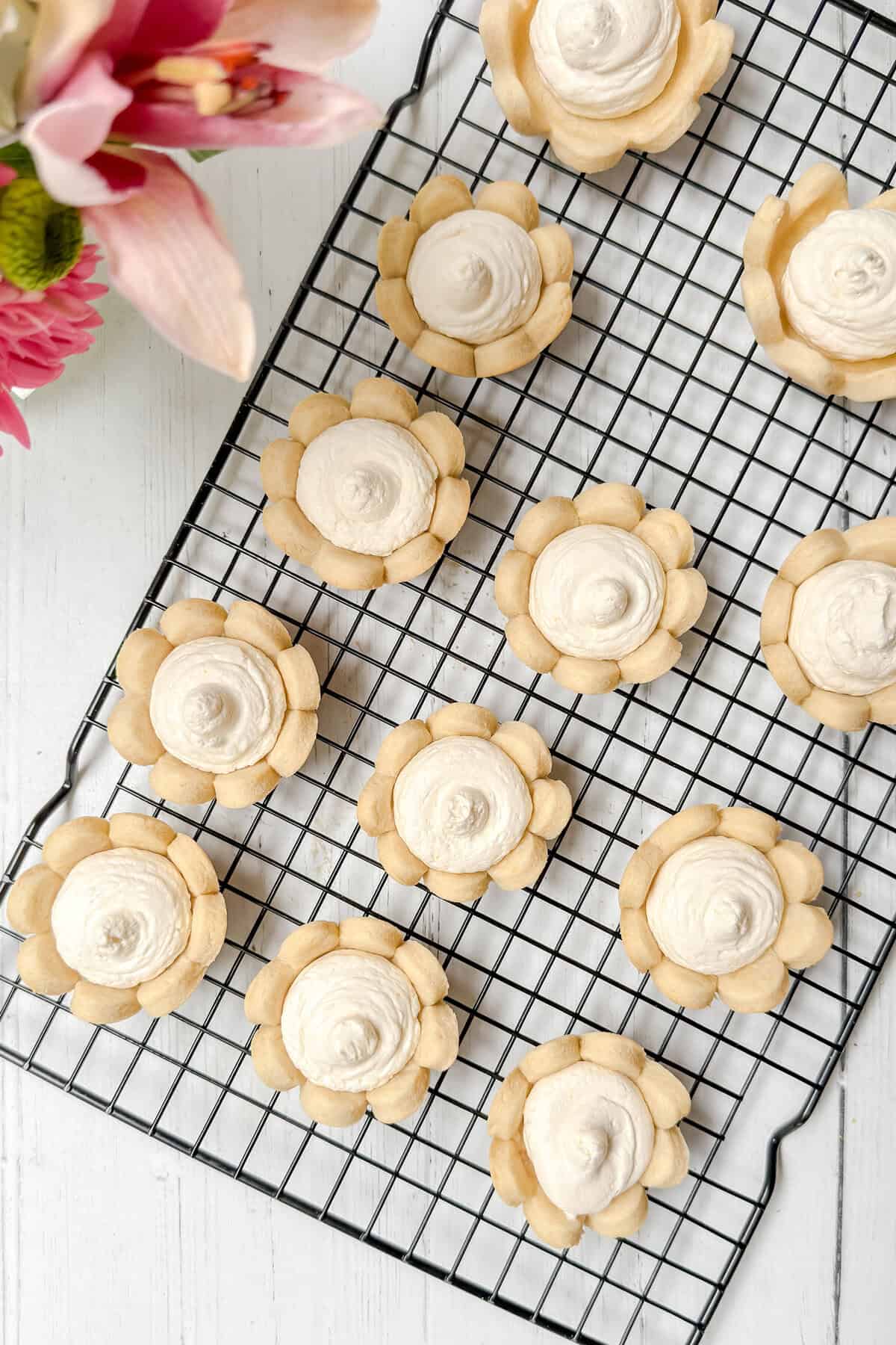 Flower-shaped sugar cookies with white frosting are arranged on a black cooling rack. Next to them, a bouquet of pink lilies and flowers sits on a white wooden surface, creating a charming scene fit for enjoying strawberry tarts.