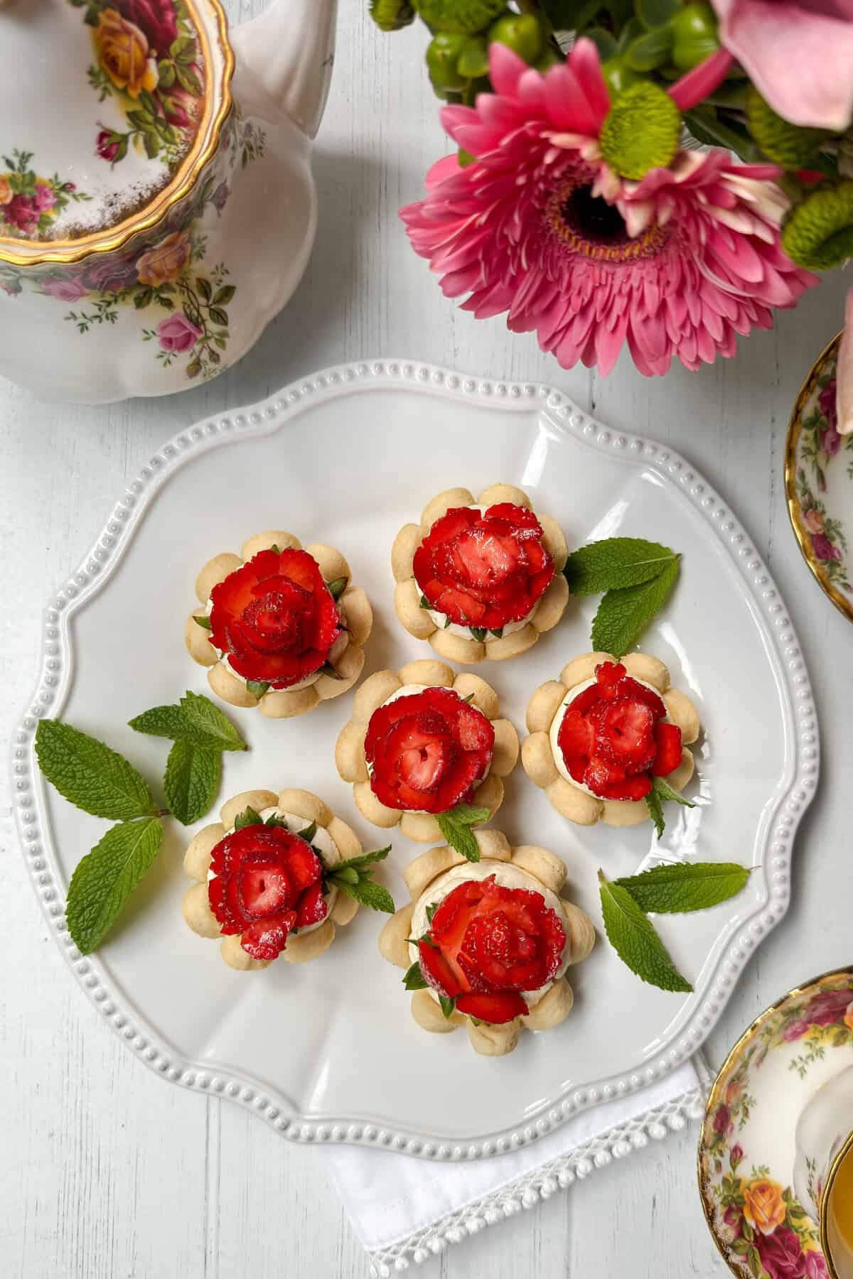 A white plate holds six flower-shaped strawberry tarts topped with mint leaves, surrounded by a teapot, floral china, and pink flowers on a white table.