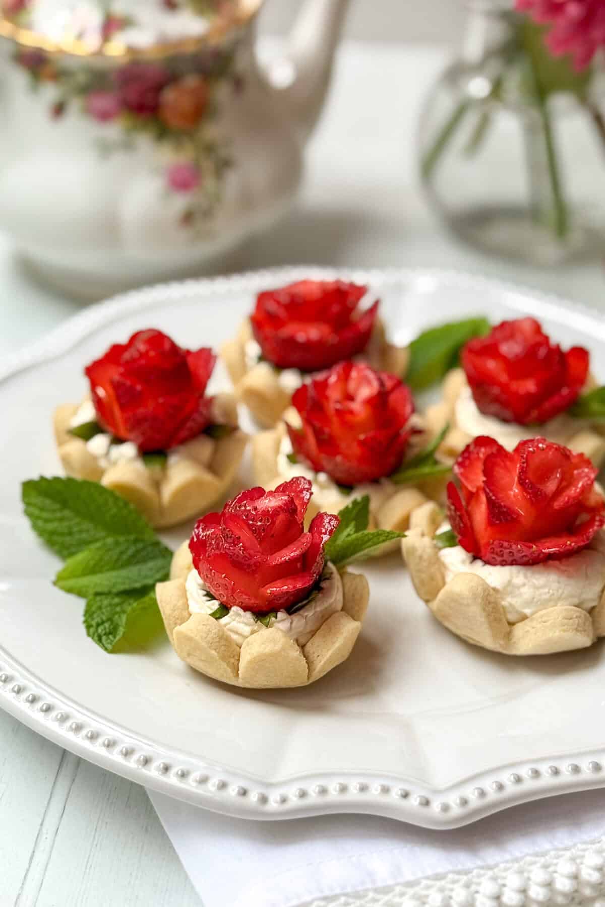 Six strawberry tarts topped with cream and strawberry slices arranged as roses are displayed on a white plate, garnished with fresh mint leaves. A floral teapot and vase are softly blurred in the background.
