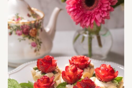 A plate of strawberry tarts sits on a white table near a teapot with floral design and a vase of pink flowers. The tarts are decorated with strawberry slices arranged like roses. Text reads "Easy Teatime Strawberry Rose Tarts.